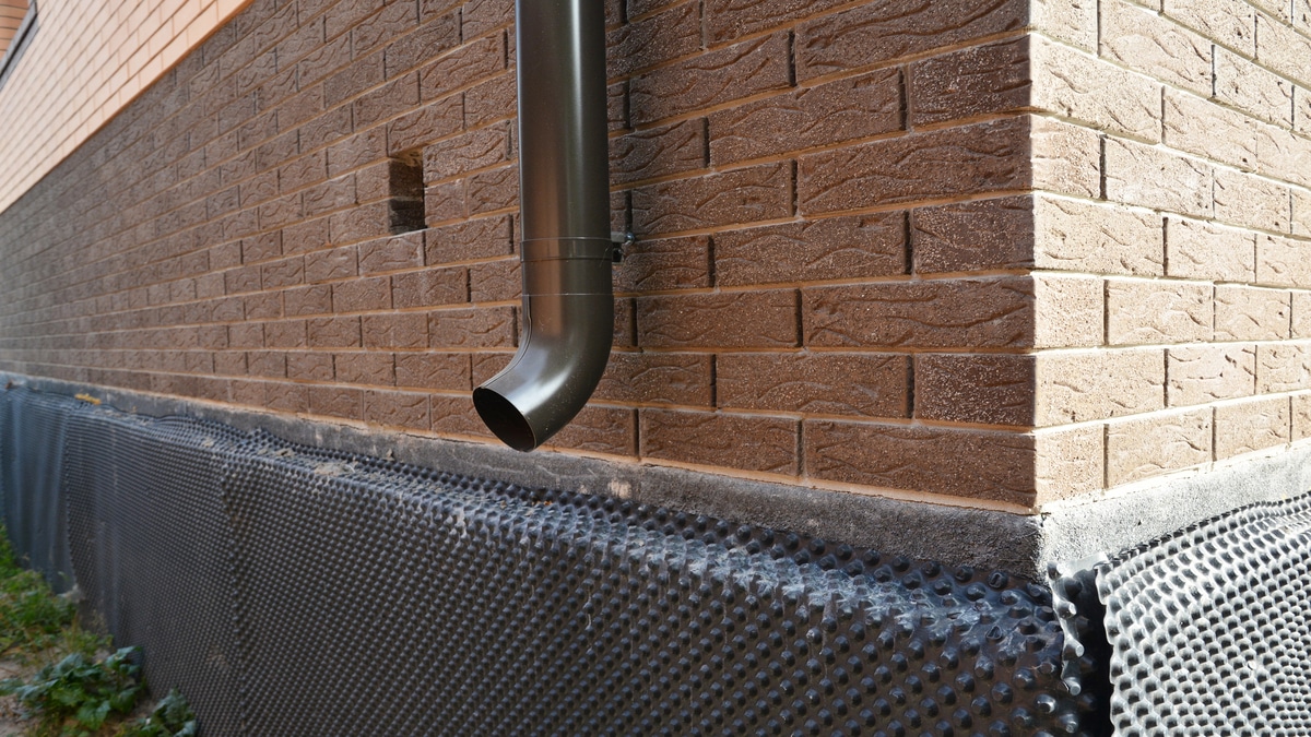 Downspout Clogs Every Fall A close-up of a metal downspout, roof gutters downspout installed near unfinished foundation, basement covered with a bitumen waterproofing membrane.
