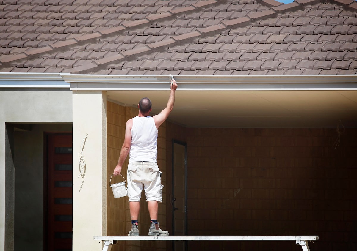 The Pros & Cons of Painting Your Gutters Man painting house exterior trim on scaffold